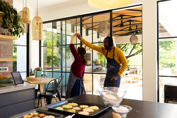 Happy diverse couple in aprons baking christmas cookies, dancing in sunny kitchen