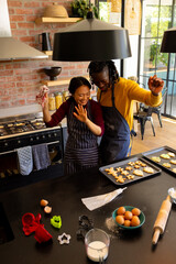 Happy diverse couple in aprons baking christmas cookies, dancing in kitchen