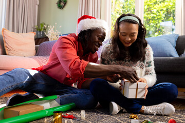 Happy diverse couple sitting on floor wrapping christmas gifts in living room - obrazy, fototapety, plakaty