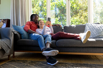 Happy diverse couple using smartphone together relaxing on couch in sunny living room