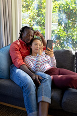 Focused diverse couple using smartphone relaxing on couch in sunny living room, copy space