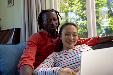 Happy diverse couple using laptop together relaxing on couch in sunny living room