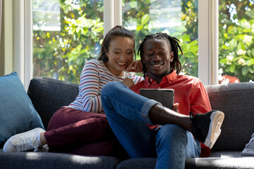 Happy diverse couple using tablet together sitting on couch smiling in sunny living room