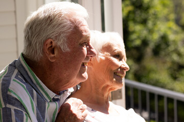 Happy caucasian senior couple embracing, smiling and looking ahead on balcony on sunny day