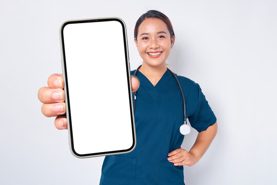 Smiling young Asian woman nurse working wearing a blue uniform showing smartphone with blank screen isolated on white background. Healthcare medicine concept