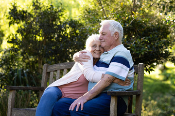 Fototapeta premium Happy caucasian senior couple sitting on bench and embracing in sunny garden, copy space