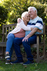 Happy caucasian senior couple sitting on bench and embracing in sunny garden, copy space