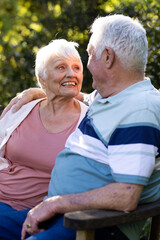 Happy caucasian senior couple sitting on bench, embracing and smiling in sunny garden, copy space