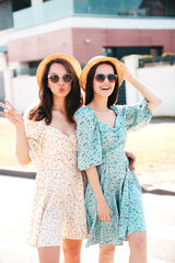 Two young beautiful smiling hipster female in trendy summer dresses  clothes. Sexy carefree women posing in the street. Positive models having fun at sunny day. Going crazy. In sunglasses and hat