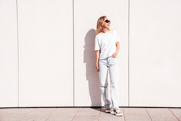 Portrait of beautiful smiling model in sunglasses. Female dressed in summer hipster white T-shirt and jeans. Posing near wall in the street. Funny and positive woman having fun outdoors
