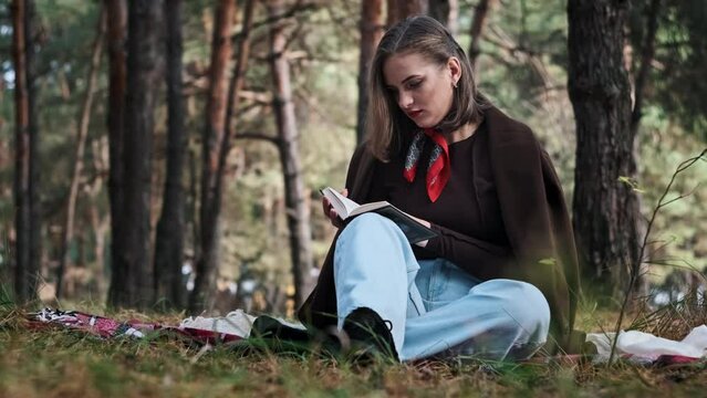 A young woman reads a book while sitting on a blanket in a pine forest. Beautiful cute girl in makeup and stylish clothes enjoys reading outdoors. Relaxing in nature. Turn the page. Slow motion. 4K.