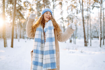 Playing with snow. Beautiful woman in a hat and scarf playing with snow, having fun in a snowy winter forest. Cheerful woman enjoying sunny frosty weather.