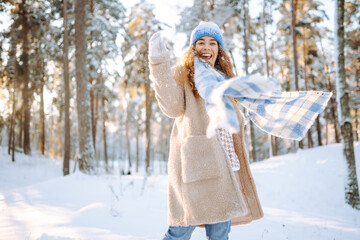 Fototapeta na wymiar Playing with snow. Beautiful woman in a hat and scarf playing with snow, having fun in a snowy winter forest. Cheerful woman enjoying sunny frosty weather.