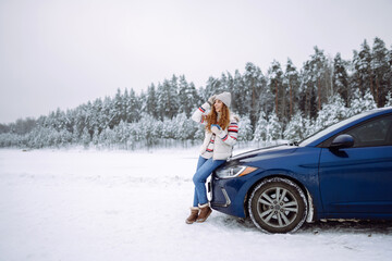 A smiling woman drinks a hot drink from a thermos while standing near her car on a winter snowy road in the forest. The concept of rest, freedom, relaxation, travel.