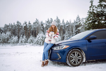 A smiling woman drinks a hot drink from a thermos while standing near her car on a winter snowy road in the forest. The concept of rest, freedom, relaxation, travel.