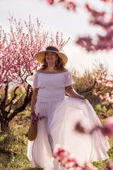 Fototapeta premium Woman blooming peach orchard. Against the backdrop of a picturesque peach orchard, a woman in a long white dress and hat enjoys a peaceful walk in the park, surrounded by the beauty of nature.