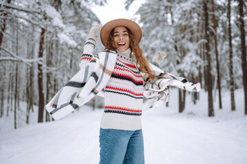 Fototapeta na wymiar Happy woman among snowy trees in winter forest, enjoying the first snow. A young woman in a hat and scarf is having fun in the winter forest. Lifestyle.