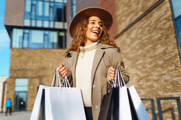 Stylish woman in a beige raincoat and hat with shopping bags in her hands. Handsome tourist near shopping center enjoying sunny weather. Shopping, shopping concept.