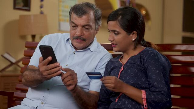 Happy 50s father with daughter making online payment for ecommerce shopping using credit card on mobile phone at home - concept of secure transaction, Banking or financial and family bonding