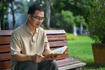 Fototapeta premium Pleased middle age man reading favorite novel while sitting on bench at public park