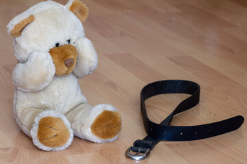Teddy bear covering eyes sits next to leather belt, violence against children concept 