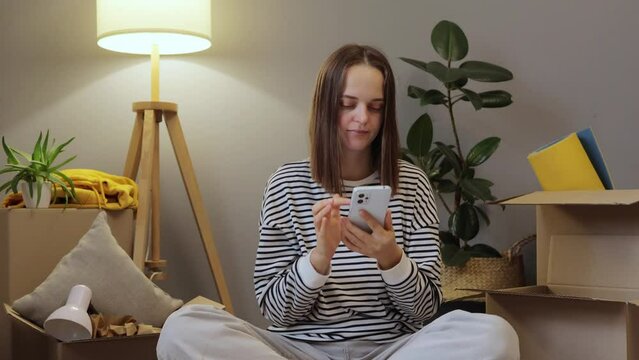 Young woman moving house. Mobile app for real estate. Female tenant using technology. Caucasian adult woman relocating sitting with her stuff in carton boxes using phone