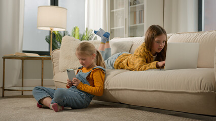 Children and modern technology two happy kids girls sisters friends play video online game together on laptop phone in cozy living room having fun playing using mobile and computer application at home