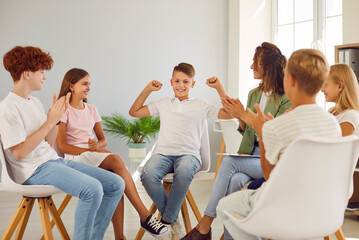 Friendly young woman conducting psychological training for a group of elementary school children sitting in a circle in the classroom applauding and looking to the boy making power winner gesture. - Powered by Adobe