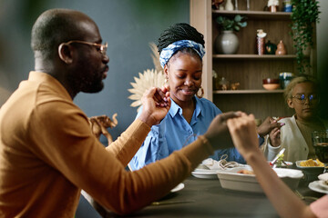 Happy young woman sitting between her husband and daughter and holding by their hands during Thanksgiving pray by festive table