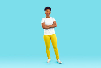Full body shot of happy beautiful young African American woman in white Tshirt, yellow pants and comfortable shoes standing with arms folded isolated on blue color background. Casual fashion concept - obrazy, fototapety, plakaty