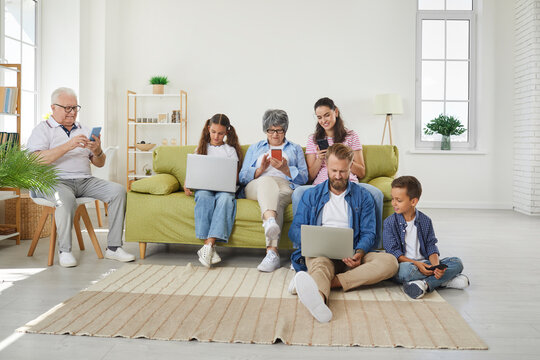Big family using different electronic devices while sitting on sofa at home. Grandparents, parents and children browsing internet, playing games or entertaining using mobile apps. Gadget addiction