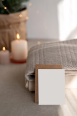 Fototapeta premium Christmas or New Year postcard or invitation template. Blank paper card mockup on table, knitted sweaters, defocused candle lights on background. Winter neutral home interior decoration