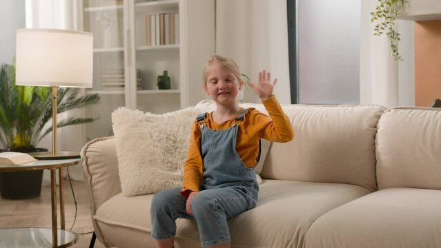 Little smiling Caucasian kid girl child daughter looking at camera waving hello greeting welcome at home couch communication with friend relative at online conference video call virtual talk concept