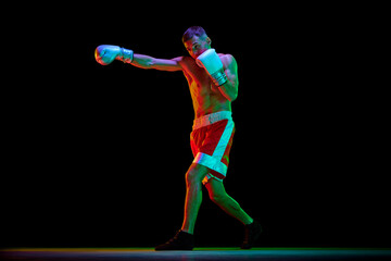 One man, professional boxer in gloves training against over black mode background in mixed neon filter, light.