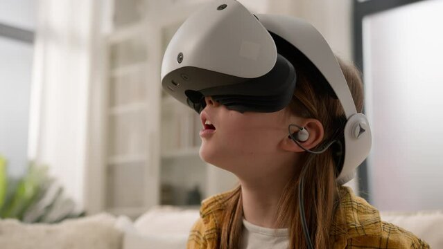 Surprised amazed Caucasian girl kid child schoolgirl playing online video game in 3D world metaverse using virtual reality helmet at home play gaming controller explore cyberspace wearing VR glasses