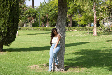Fototapeta premium Beautiful Spanish brunette woman with long curly hair and big slit eyes leans her back against the trunk of a tree. The woman is smiling and happy and enjoying the day in the city of Seville in Spain.