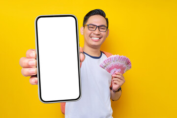 Smiling young Asian student man in casual clothes and glasses backpack holding smartphone blank screen and cash money isolated on yellow background. Education in High School University College concept - obrazy, fototapety, plakaty