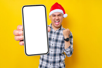 Excited young Asian man in plaid shirt wearing Christmas hat holding mobile phone with blank...