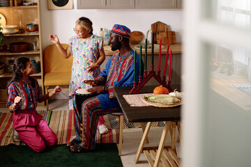 Happy young African American man beating drum and looking at his daughters in national costumes...