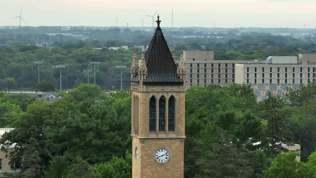 Iconic Campanile tower at Iowa State University, set against lush greenery, with distant buildings and wind turbines. Aerial shot with zoom lens.