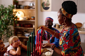 Side view of young African American woman in national dress and accessories burning candles symbolizing seven kwanzaa principles - obrazy, fototapety, plakaty