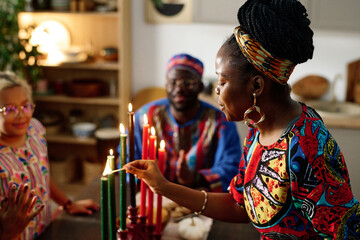 Side view of young African American woman in ethnic apparel burning candles symbolizing seven main principles of national culture - obrazy, fototapety, plakaty