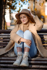 Beautiful adult blonde woman in a hat and coat sits on a bench in an autumn park and has fun