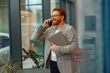 Bearded businessman in eyeglasses with laptop is talking phone standing in modern coworking space