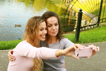 Fototapeta premium mature mother and adult daughter are doing selfie by mobile phone near lake in summer park