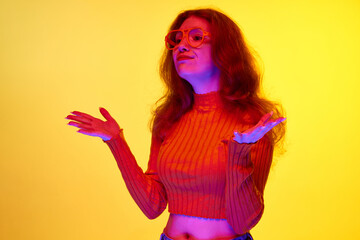 Young girl in casual clothes wearing glasses, spreading hands against yellow studio background in neon light. Concept of youth, human emotions, facial expression, lifestyle