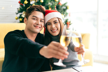 Young happy Asian man smile and laughs in the back at his girlfriend wearing a Santa Claus hat while drinking champagne at home with a Christmas tree in the background.