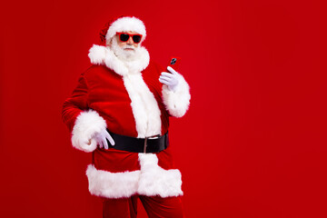 Portrait of good mood smart santa claus in trendy sunglass look at smartphone hand on waist chatting isolated on red color background