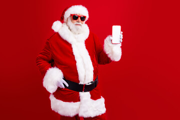 Portrait of intelligent cool santa claus in sunglass demonstrate smartphone touchscreen empty space isolated on red color background