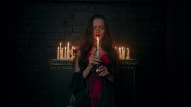 A young beautiful witch is fortune-telling by candlelight in a dark room in a red corset. A beautiful brunette in a dark room fortune-telling on a candle